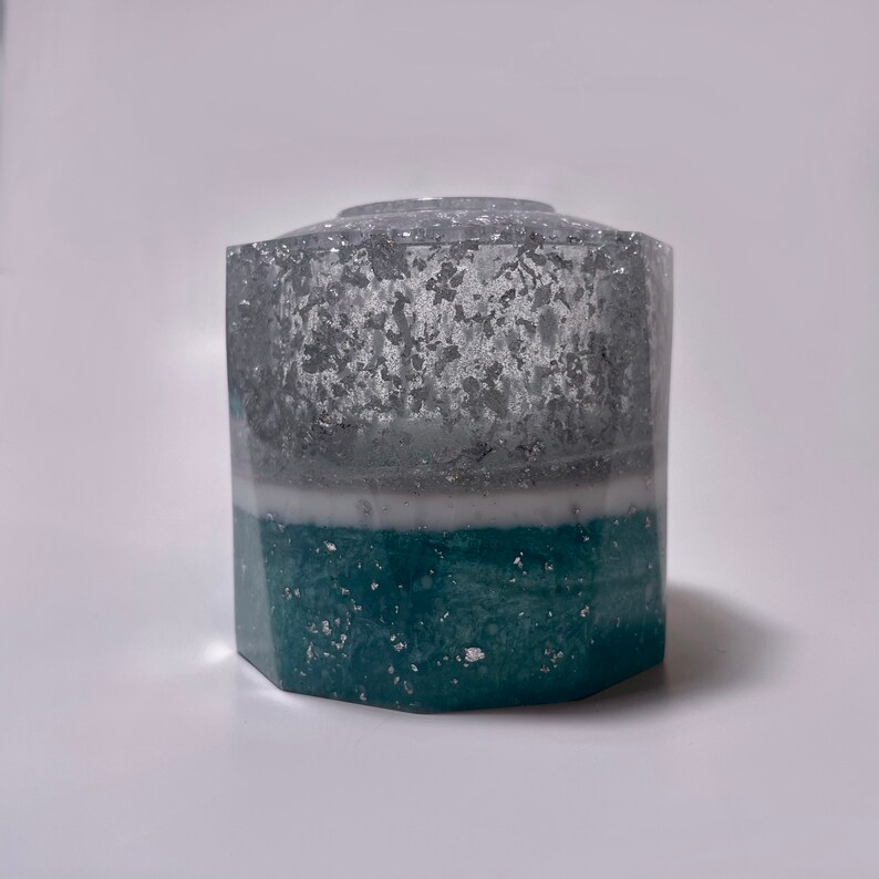 May include: A hexagonal, clear glass vase with a teal and silver glitter design. The vase has a white layer in the middle.
