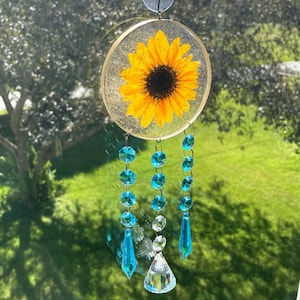 May include: A round, clear resin suncatcher with a pressed sunflower inside. The suncatcher is hanging from a suction cup and has three strands of turquoise crystal beads hanging from the bottom.