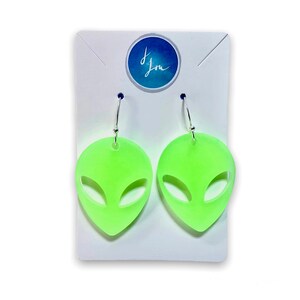 May include: A pair of green alien face earrings with silver hooks. The earrings are on a white card with the text "J. Lou" on it.