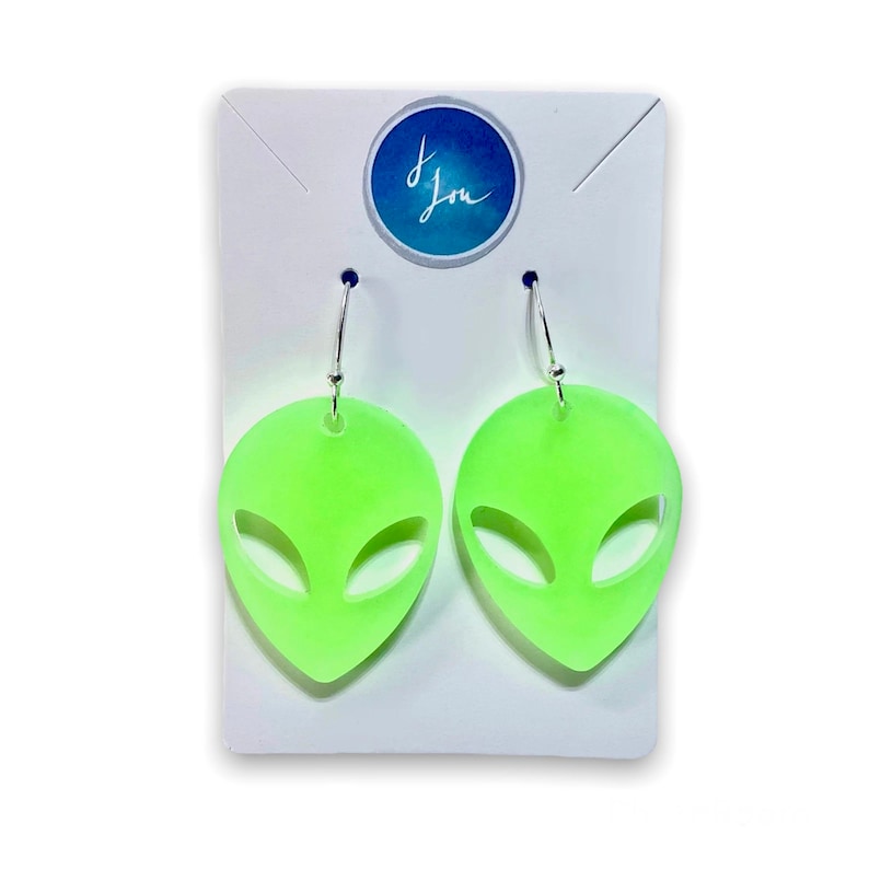 May include: A pair of green alien face earrings with silver hooks. The earrings are on a white card with the text "J. Lou" on it.