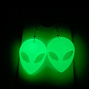 May include: A pair of green glow-in-the-dark alien head earrings. The earrings are made of a smooth, textured material and have a silver hoop.