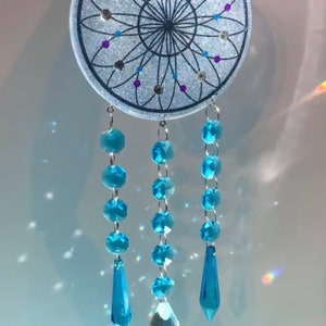 May include: A white dreamcatcher with a black web design and blue and purple glitter accents. The dreamcatcher has three strands of blue crystal beads with teardrop-shaped crystal pendants.