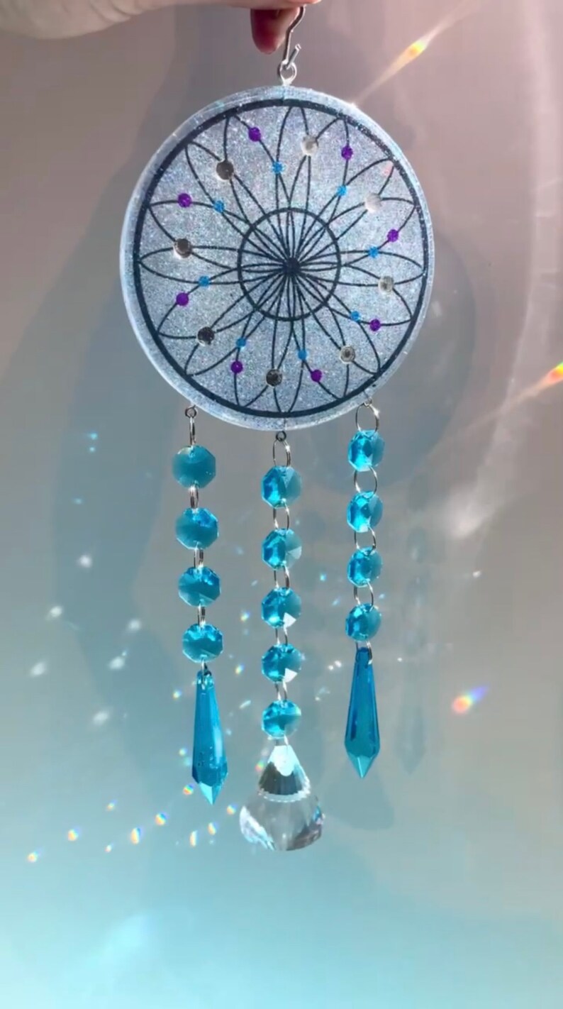 May include: A white dreamcatcher with a black web design and blue and purple glitter accents. The dreamcatcher has three strands of blue crystal beads with teardrop-shaped crystal pendants.