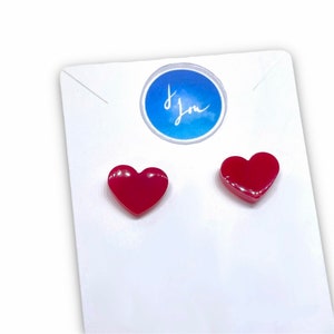 Op de afbeelding: Een paar rode hartvormige oorbel studs. De oorbellen liggen op een witte kaart met de woorden "Ik hou van je" op een blauwe cirkel.