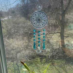 May include: A silver dreamcatcher with a black and white geometric design and turquoise crystal beads hanging from it. The dreamcatcher is hanging from a window with a green plant in the foreground.