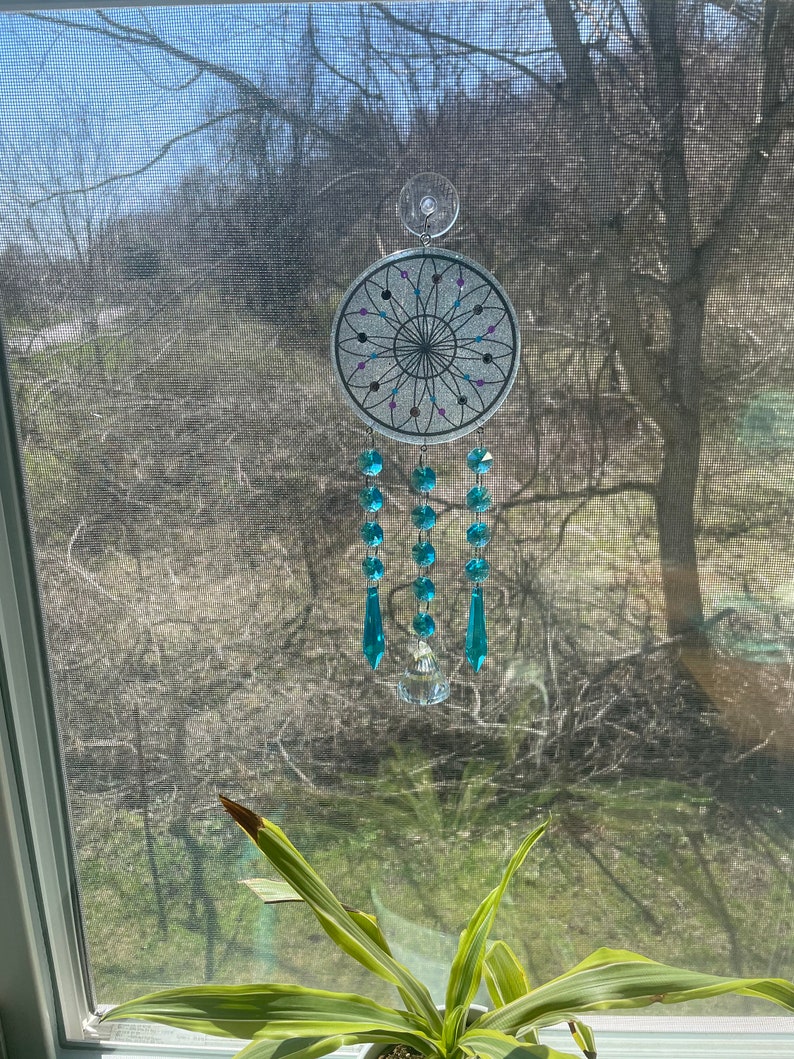 May include: A silver dreamcatcher with a black and white geometric design and turquoise crystal beads hanging from it. The dreamcatcher is hanging from a window with a green plant in the foreground.