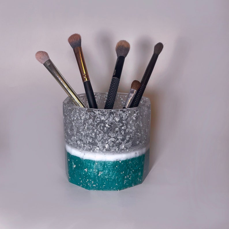 May include: A teal and silver makeup brush holder with five brushes inside. The holder is made of resin and has a textured, glittery finish.