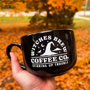 May include: Black ceramic coffee mug with the text "Witches Brew Coffee Co. Stirring Up Trouble" in white. The design includes a witch's hat, moon, and stars. The mug has a curved handle.