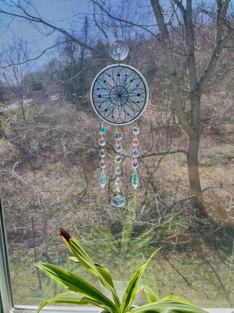 May include: A white dreamcatcher with iridescent beads and crystal accents hanging from a window. The dreamcatcher has a black and white geometric design with purple and blue accents.
