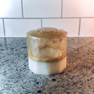 May include: A white and gold hexagonal resin container with gold glitter embedded in the clear resin. The container has a white base and a clear top with gold glitter.