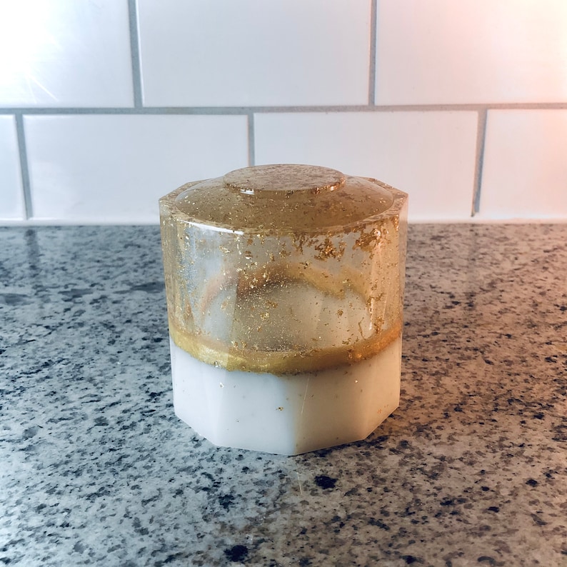 May include: A white and gold hexagonal resin container with gold glitter embedded in the clear resin. The container has a white base and a clear top with gold glitter.