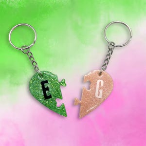 Puede incluir: Dos piezas de rompecabezas en forma de corazón con purpurina, una verde y otra rosa, con las letras "E" y "G" en ellas. Las piezas están unidas a llaveros plateados.