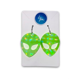 May include: A pair of green alien head-shaped earrings with iridescent accents. The earrings are on a white card with a blue circle that says "J. Lou".
