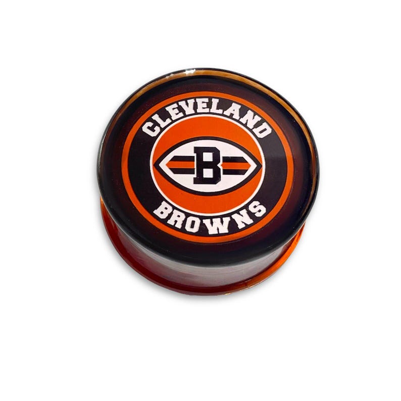 Puede incluir: Un tap&oacute;n redondo de vidrio, naranja y negro, con el logotipo de los Cleveland Browns. El logotipo presenta una "B" blanca dentro de una forma de bal&oacute;n de f&uacute;tbol naranja con rayas negras.