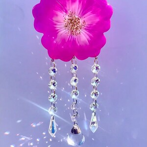 May include: A pink resin flower with a white centre and gold glitter accents hangs from three strands of crystal beads with teardrop-shaped crystal pendants.