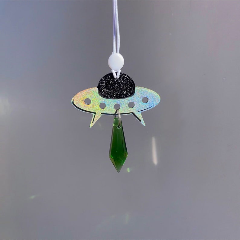 May include: A silver iridescent UFO charm with a black glitter top and a dangling green crystal pendant. The charm is attached to a white string.