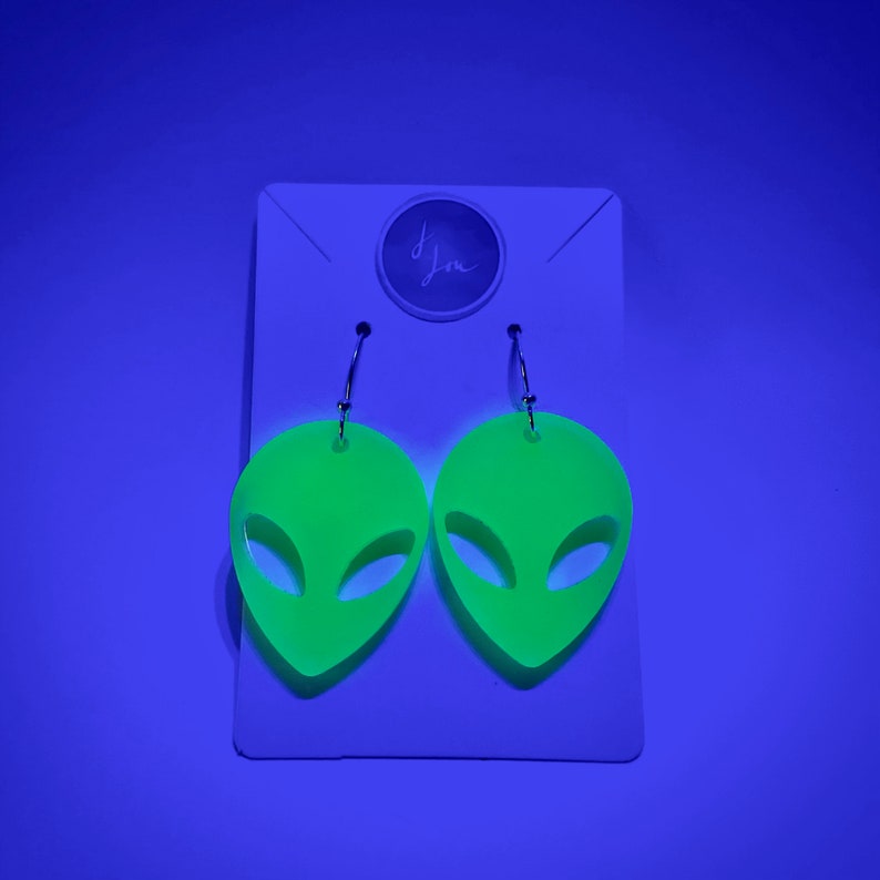 May include: A pair of neon green alien-shaped earrings with silver hooks. The earrings are on a white card with the word "Im" written in black.