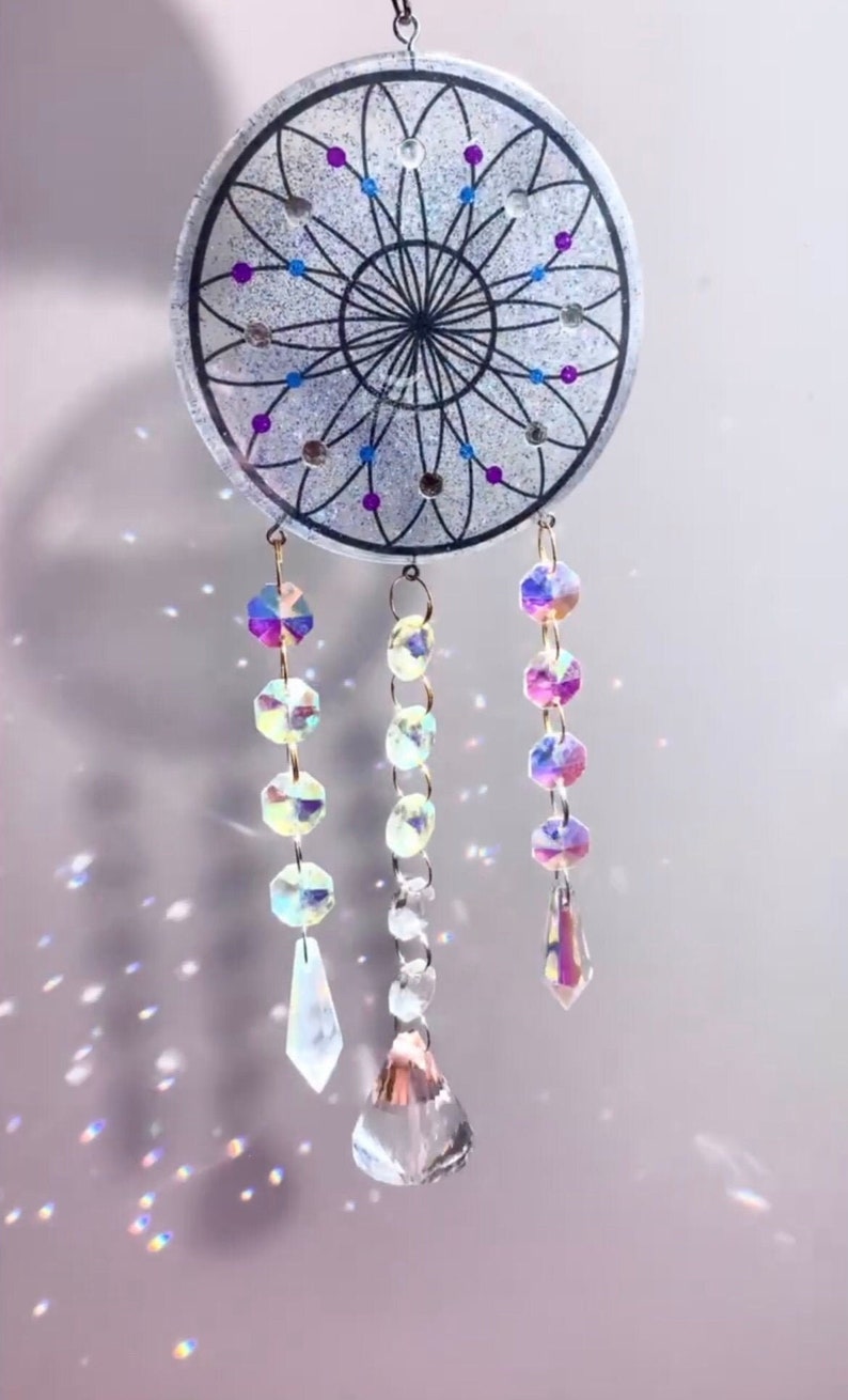 May include: A silver dreamcatcher with a black web design and purple and blue accents. The dreamcatcher has three strands of iridescent crystal beads hanging from it.