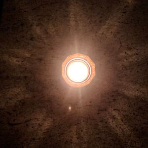 May include: A round, gold-colored light fixture with a white light bulb shining brightly. The light fixture is mounted on a brown ceiling.