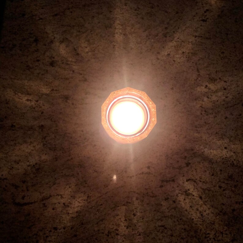 May include: A round, gold-colored light fixture with a white light bulb shining brightly. The light fixture is mounted on a brown ceiling.