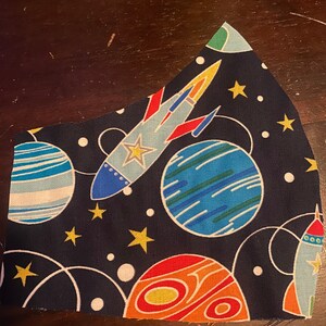 Op de afbeelding: Een donkerblauw stoffen mondkapje met een ruimtethema print met planeten, sterren en raketten. De planeten zijn blauw, oranje en groen, en de raketten zijn rood, geel en blauw.