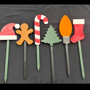 May include: A collection of Christmas-themed yard stakes. Includes a red Santa hat, gingerbread man, candy cane, Christmas tree, orange light bulb, and red stocking. Each stake has a decorative top and a pointed bottom for easy placement.