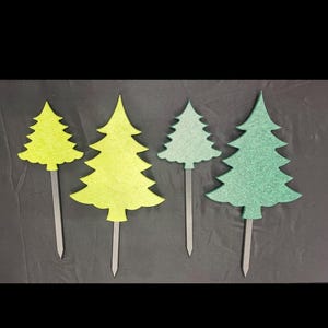 May include: Four decorative tree-shaped toppers in varying shades of green. Each tree has a textured appearance and is attached to a silver-colored stake. The toppers are arranged against a dark background.