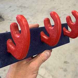 May include: A wooden wall-mounted coat rack with three red antler-shaped hooks attached to a dark blue rectangular base. The hooks are curved and have a textured surface, providing a rustic and functional design for hanging items.