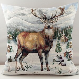 Può includere: Cuscino quadrato decorativo con un design arazzo dettagliato di un maestoso cervo in un paesaggio invernale innevato. Il cuscino mostra un cervo marrone con grandi corna, sullo sfondo di montagne innevate, alberi e piccoli edifici.