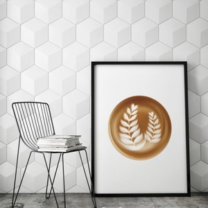 May include: A framed print featuring a latte art design with two leaf-like patterns in a brown cup. The print is displayed against a white geometric wall. A black wire chair and a stack of books are in the foreground.