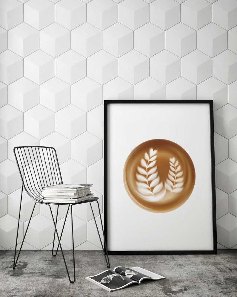 May include: A framed print featuring a latte art design with two leaf-like patterns in a brown cup. The print is displayed against a white geometric wall. A black wire chair and a stack of books are in the foreground.