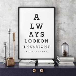Puede incluir: Un cartel enmarcado con el texto "ALWAYS LOOK ON THE BRIGHT SIDE OF LIFE" en negro. El cartel está sobre un mueble azul oscuro, junto a candelabros, libros y una campana de cristal con una piña.