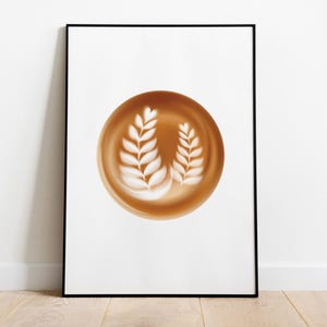 May include: A framed print featuring a coffee art design. The artwork showcases a brown coffee-colored circle with two white leaf-like designs. The frame is black, and the background is white. The print is displayed on a wooden floor.