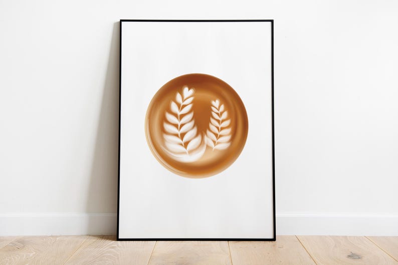 May include: A framed print featuring a coffee art design. The artwork showcases a brown coffee-colored circle with two white leaf-like designs. The frame is black, and the background is white. The print is displayed on a wooden floor.