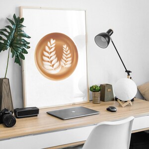 May include: A framed print featuring a latte art design of two white leaf patterns on a brown background. The print is displayed on a wooden desk with a laptop, camera, and desk lamp.