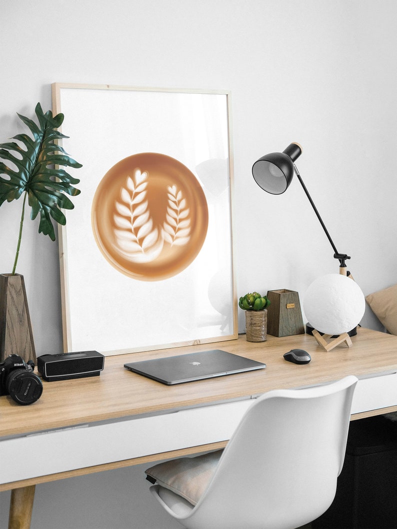 May include: A framed print featuring a latte art design of two white leaf patterns on a brown background. The print is displayed on a wooden desk with a laptop, camera, and desk lamp.
