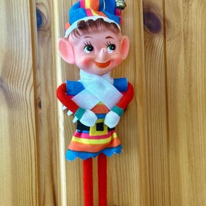 Könnte beinhalten: Eine hängende Elfenpuppe mit einer spitzen Mütze und einer kleinen Glocke, einem bemalten Gesicht mit großen grünen Augen und einem bunten, gestreiften Outfit. Die Puppe hat lange rote Filzbeine, schwarze spitze Schuhe und weiße Handschuhe.