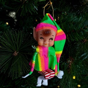 Op de afbeelding: Een hangend elf-ornament met een puntmuts en tuniek in neon groene, roze en oranje strepen. De elf heeft een gevormd plastic gezicht met geschilderde gelaatstrekken, bruin haar en puntige oren. Het draagt rood-wit gestreepte leggings en witte laarzen.
