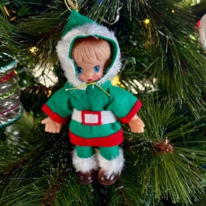 Op de afbeelding: Een kleine elfenpop als ornament met een gevormd gezicht, blauwe ogen en bruin haar. De pop draagt een groene vilten tuniek met rode randen, een witte riem en een groene broek. Een capuchon met witte imitatiebontrand bedekt het hoofd en de broekspijpen.