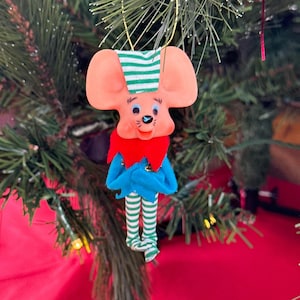 May include: A hanging Christmas ornament shaped like a mouse with a peach-coloured face, wearing a green and white striped hat, a red collar, a blue jacket, and green and white striped legs. The mouse has black eyes, a black nose, and whiskers.