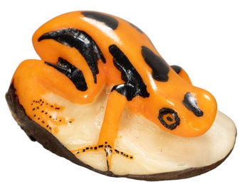 May include: A small, orange and black poison dart frog figurine perched on a white, textured base. The frog has a black stripe running down its back and black spots on its body.