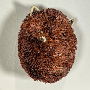 May include: A brown, spherical decorative object resembling a hedgehog. The body is covered in short, textured, brown pieces, with small, white-lined ears and a small, white eye visible. The object is set against a plain white background.