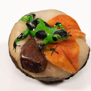 May include: A green and black frog figurine perched on a brown and orange leaf sculpture. The sculpture is made of a light brown material and has a rough texture.