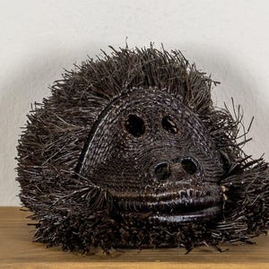 May include: A dark brown, woven sculpture of a gorilla's head. The sculpture has detailed facial features, including eyes, a nose, and a mouth. The gorilla's head is displayed on a wooden surface.