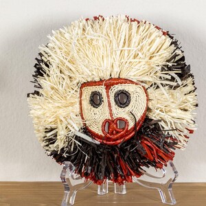 May include: A round, woven mask with a face design. The mask features a red and cream-colored face with dark eye and mouth details. The mask is surrounded by a fringe of cream, black, and red fibers, and is displayed on a clear stand.