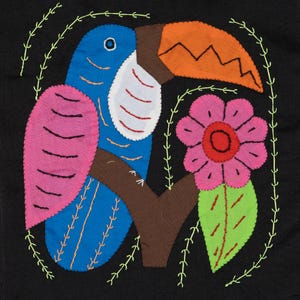 May include: A colorful toucan with a pink wing and orange beak perched on a brown branch with a pink flower and green leaf. The toucan is embroidered on a black fabric background.