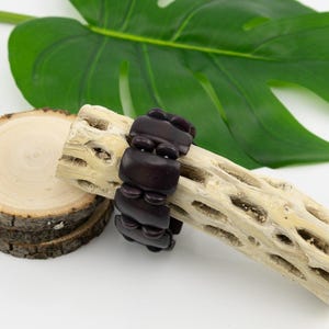Puede incluir: Una pulsera de cuentas marrón oscuro descansa sobre un trozo de madera clara y texturizada. Una pila de rodajas de madera y una gran hoja verde están en el fondo. La pulsera está hecha de cuentas rectangulares y redondas.