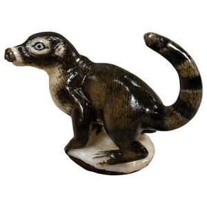May include: A small ceramic figurine of a ring-tailed animal, likely a coati or lemur, featuring a dark brown and cream-colored coat. The creature stands on a white base with its long, striped tail curved upward in a distinct arching position.