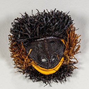 May include: A handcrafted mask with a monkey-like face. The mask features a black woven face with eye and nose openings, a yellow mouth, and a fringe of black and brown material around the face. The mask is likely for costume or decorative purposes.