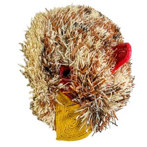 May include: A handcrafted mask with a textured, shaggy appearance. The mask features a mix of beige, brown, and black materials, with red accents for the eyes and ear. A yellow, woven section forms the lower part of the face.